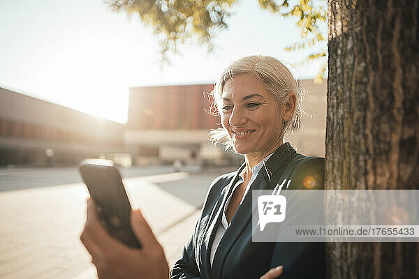 Smiling businesswoman using smart phone by tree trunk at office park on sunny day