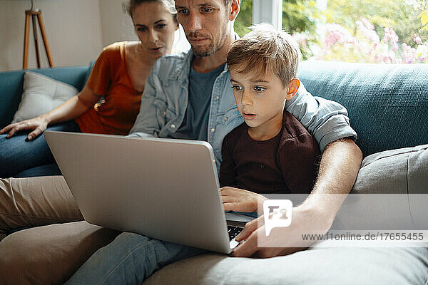 Father and son sharing laptop sitting by woman on sofa at home