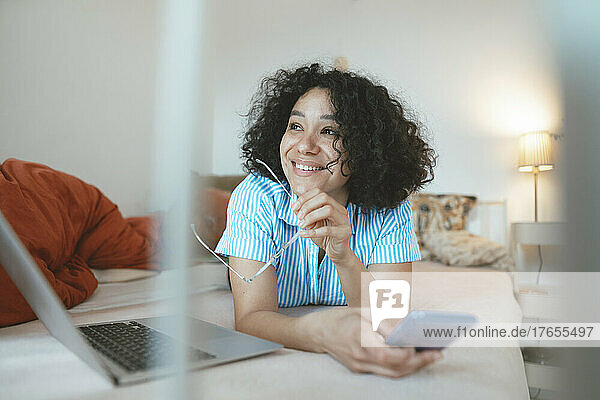 Happy woman with laptop lying on bed holding mobile phone at home