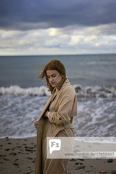 Young woman wearing trench coat standing in front of Baltic Sea at sunset