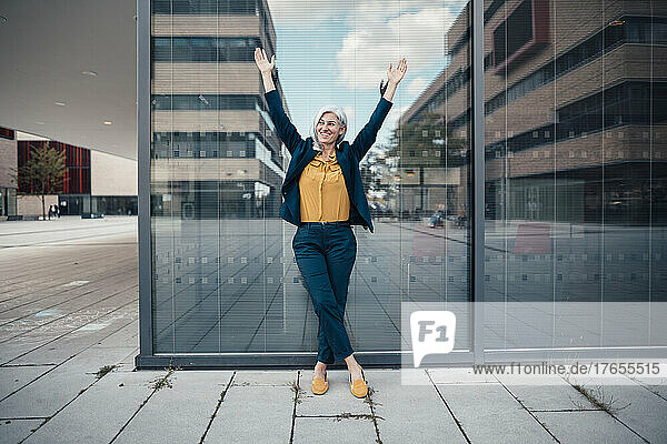 Happy businesswoman with arms raised standing in front of glass wall at office park