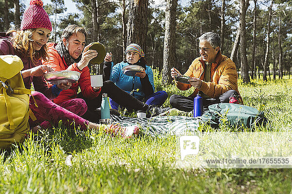 Smiling men and women eating food sitting in forest on sunny day