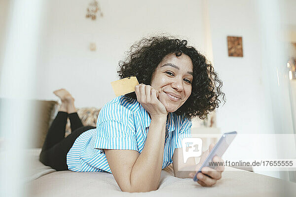 Smiling woman with credit card using smart phone on bed at home