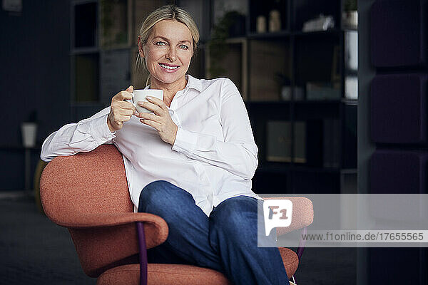 Lächelnde Geschäftsfrau mit Kaffeetasse sitzt auf einem Stuhl im Büro