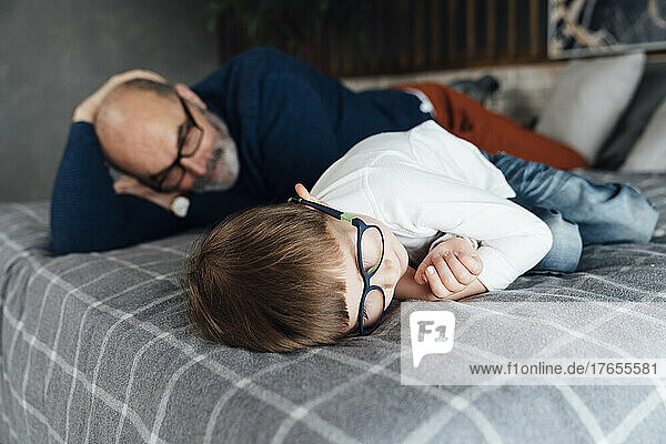 Boy wearing eyeglasses lying by grandfather on bed at home