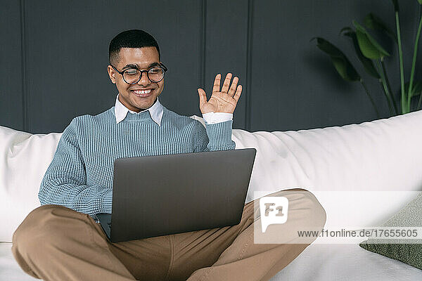 Happy freelancer waving hand on video call through laptop at home