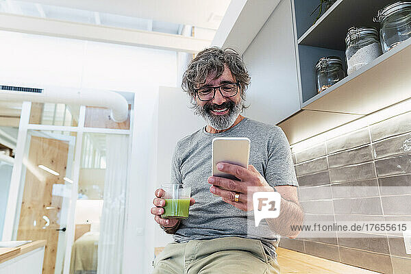 Man sitting on kitchen counter with smoothie and smart phone in kitchen at home