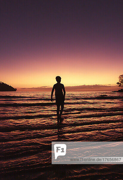 Silhouette of boy standing in shallow water on a beach at sunset.