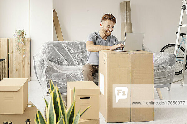 A happy young man using his laptop on a cardboard box in his new home