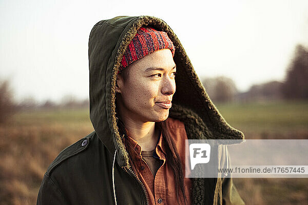 Non-binary person smiles in afternoon sun outdoors in field with hood