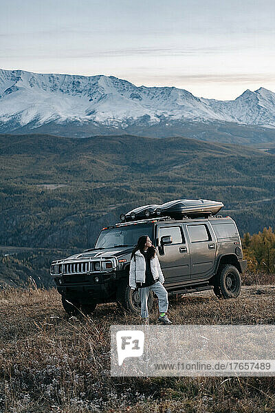 A girl stands next to a black SUV background of a mountain range