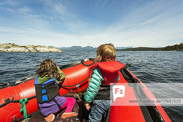 Two young kids sit at bow of rubber raft looking at rocky island
