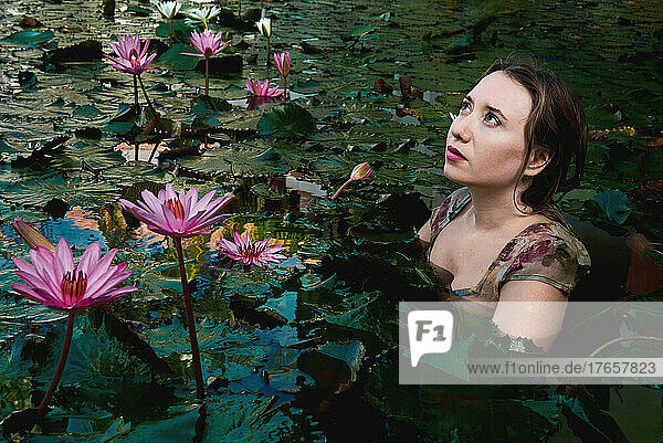 a girl sits in a lake with lotuses looking at the sky with hope