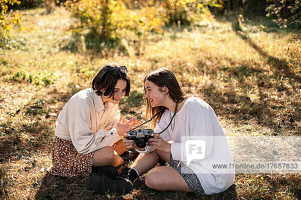Happy female friends with camera sitting on grass in park in autumn