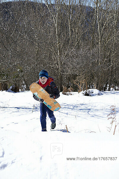 Cheerful teen male running on snow while holding a surf Board
