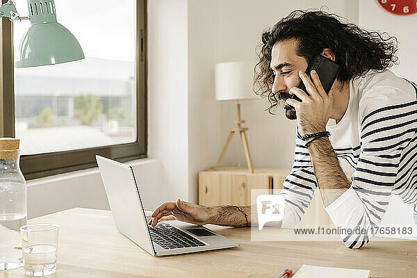 Cheerful man talking on phone with his laptop at home office.