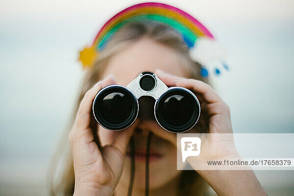 Girl with blond hair and rainbow headband looks through binoculars