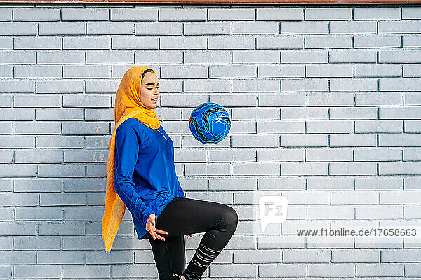 Ethnic woman kicking ball with knee near brick wall