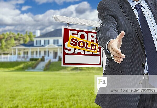 Immobilienmakler greift zum Handschlag mit Verkaufsschild und neuem Haus im Rücken