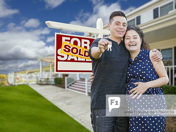 Glückliches hispanisches Paar vor neuem Haus und verkauftem Immobilienschild  das seine Hausschlüssel zeigt
