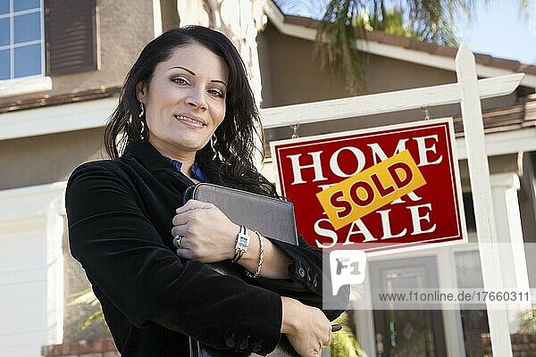 Stolze  attraktive hispanische Frau vor einem verkauften Immobilienschild und einem neuen Haus