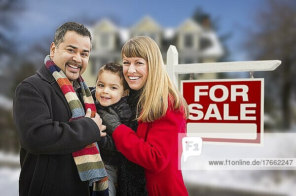 Warm gekleidete junge gemischtrassige Familie vor einem zu verkaufenden Immobilienschild und einem Haus mit Schnee auf dem Boden