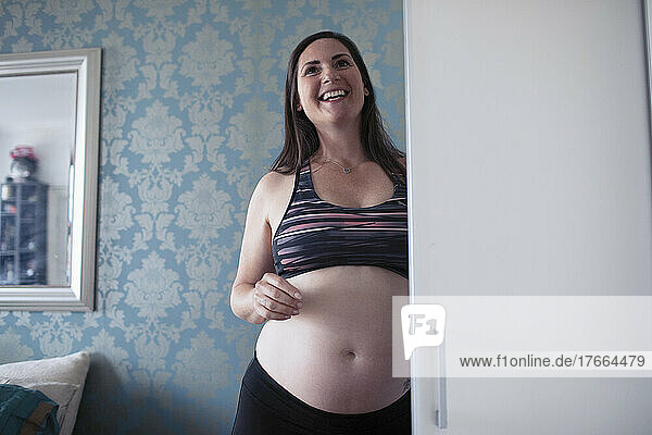 Glückliche schwangere Frau im Sport-BH am Schlafzimmerschrank