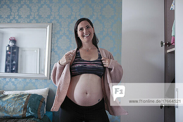 Portrait happy pregnant woman putting on sweater at closet