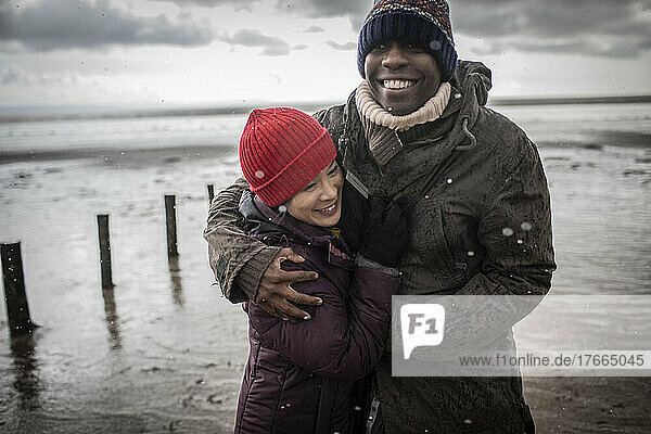 Glückliches Paar in warmer Kleidung  das sich am Winterstrand umarmt