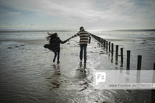 Paar hält sich an den Händen und läuft am sonnigen  nassen Winterstrand