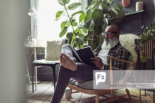 Barfuß reifer Mann mit Bart liest Buch zu Hause
