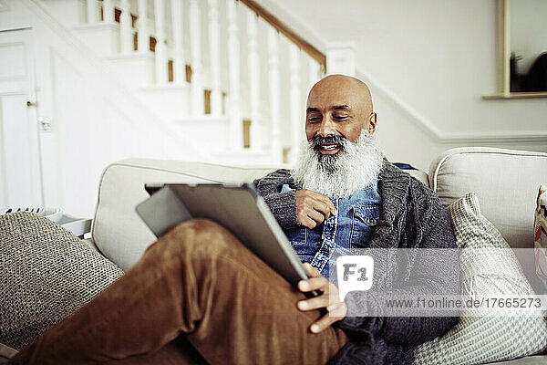 Älterer Mann mit Bart  der auf dem Wohnzimmersofa ein digitales Tablet benutzt