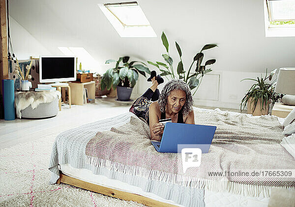Ältere Frau mit Kreditkarte bezahlt Rechnungen am Laptop auf dem Bett