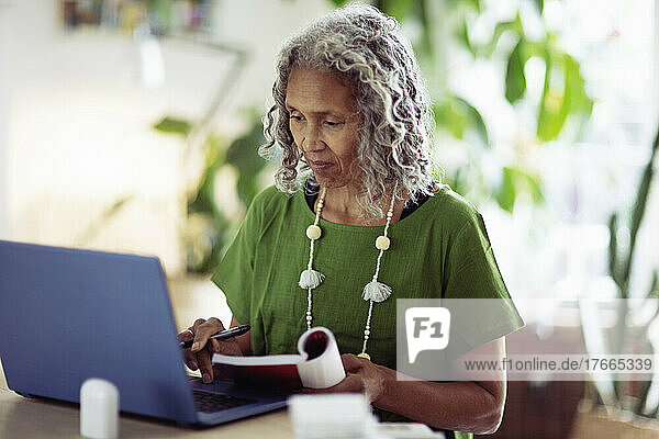 Ältere Frau mit Buch arbeitet zu Hause am Laptop
