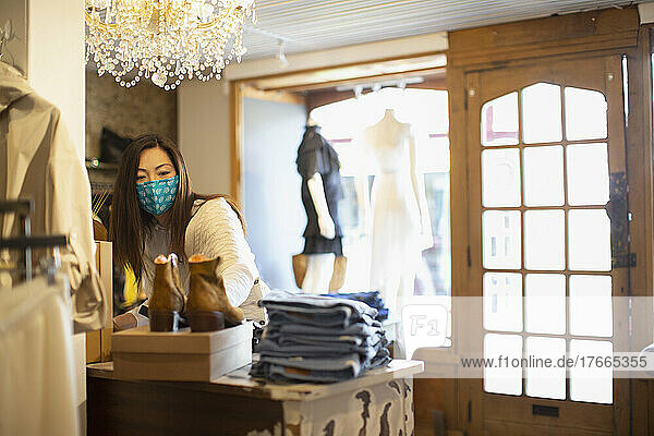 Weibliche Ladenbesitzerin mit Gesichtsmaske arrangiert die Auslage in einer Bekleidungsboutique