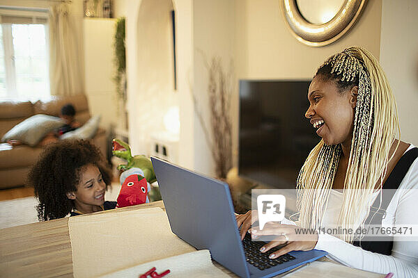 Glückliche Mutter arbeitet am Laptop und sieht ihrer Tochter beim Spielen zu