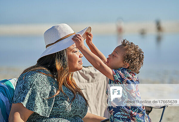 Curious son with Down Syndrome lifting mother's hat on beach