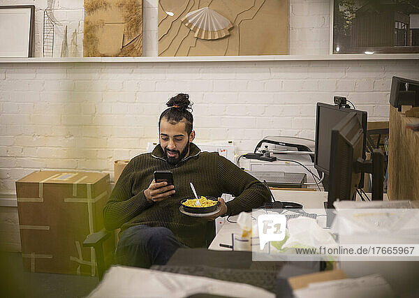 Young businessman eating lunch and using smart phone in office