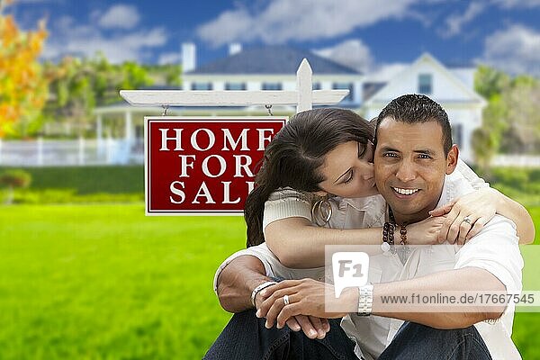 Junges glückliches hispanisches junges Paar vor ihrem neuen Haus und zum Verkauf stehenden Immobilienschild