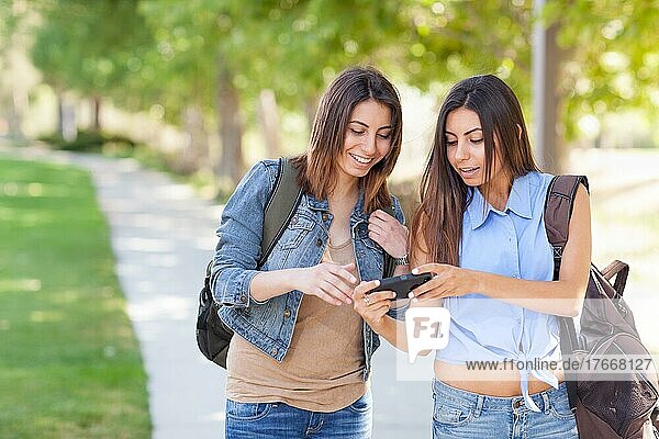Zwei schöne junge ethnische Zwillingsschwestern mit Rucksäcken  die draußen ein Smartphone benutzen