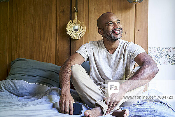 Smiling man holding smart phone while sitting in bedroom