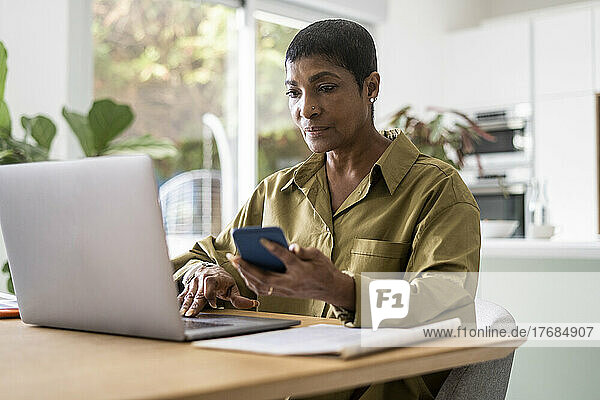 Mature woman holding smart phone and using laptop