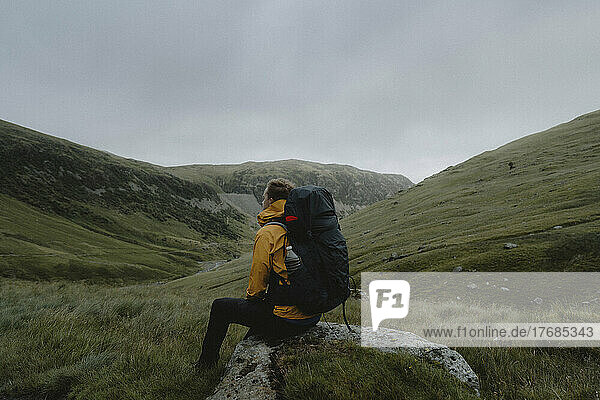 Male hiker with backpack resting on rock in grassy remote landscape  Red Tarn  England