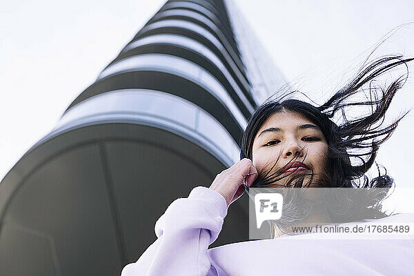 Junge Frau mit zerzausten Haaren steht vor einem Wolkenkratzer