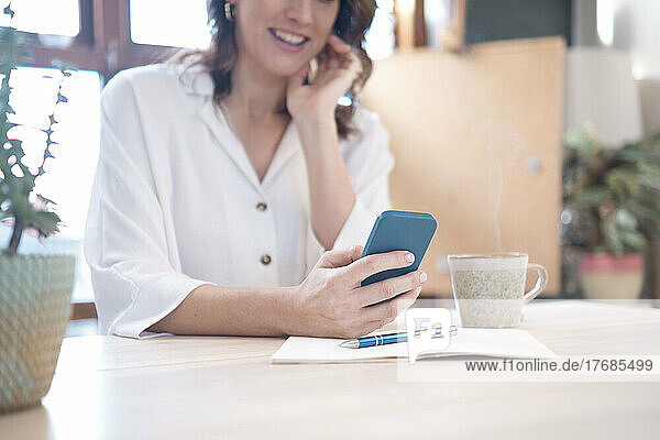 Lächelnde Frau mit Smartphone und Tagebuch sitzt zu Hause am Tisch