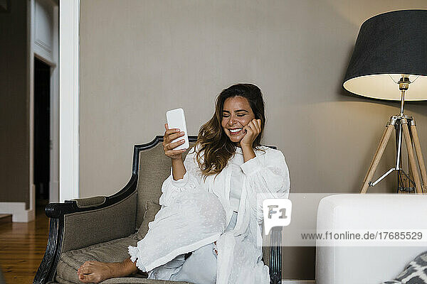 Glückliche Frau mit Smartphone sitzt zu Hause im Sessel