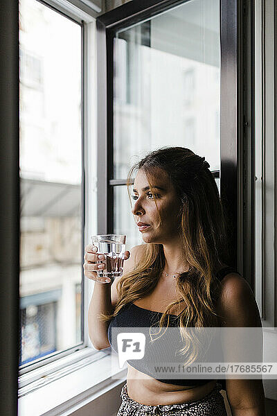 Junge Frau hält ein Glas Wasser in der Hand und schaut zu Hause durch das Fenster
