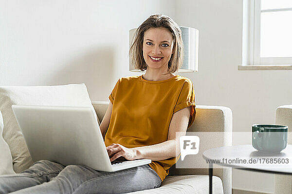 Lächelnde Frau mit Laptop sitzt auf dem Sofa im Wohnzimmer