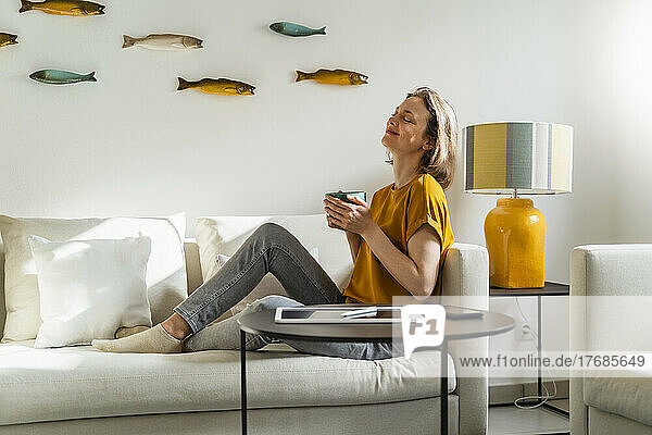 Smiling woman with eyes closed holding coffee mug sitting on sofa at home