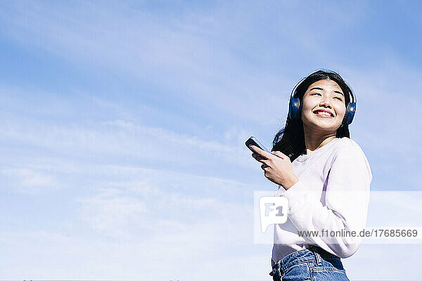 Lächelnde Frau mit Kopfhörern und Smartphone an einem sonnigen Tag
